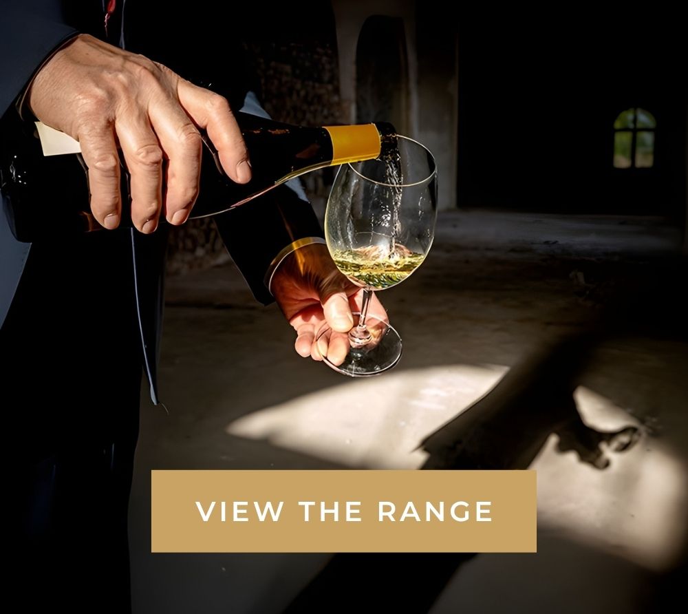 Person pouring white wine from a bottle into a glass in a dimly lit room with a window in the background.