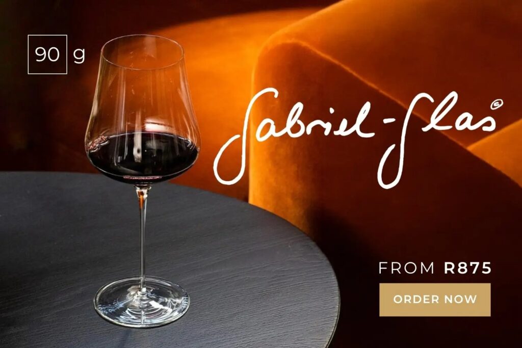 Elegant wine glass with red wine on a dark table, orange velvet chair in background, Gabriel-Glas logo, and order details.