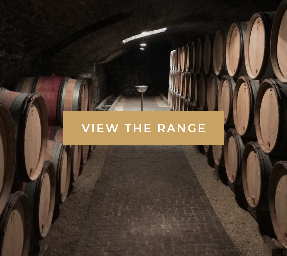 Wine barrels line a dim stone cellar, with a central brick walkway and “VIEW THE RANGE” text overlay.